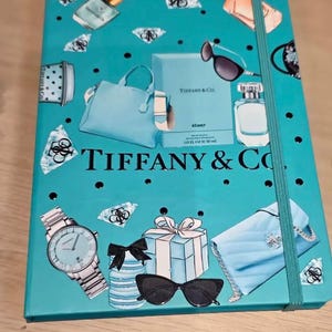 以下が含まれることがあります： 「TIFFANY & Co.」の文字と、時計、サングラス、香水、ハンドバッグ、ギフトボックスのイラストが描かれた、ターコイズブルーのノート。 ターコイズブルーのゴムバンドが付いています。