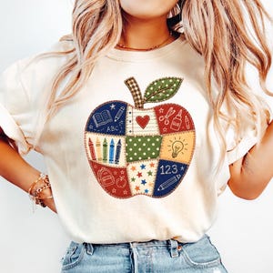 Könnte beinhalten: Ein cremefarbenes T-Shirt mit einer großen, zentralen apfelförmigen Grafik, die aus verschiedenen quadratischen Patchwork-Mustern besteht. Das Design zeigt schulbezogene Symbole wie Buntstifte, einen Rucksack, eine Glühbirne, Schere, Kleber sowie ABC und 123.