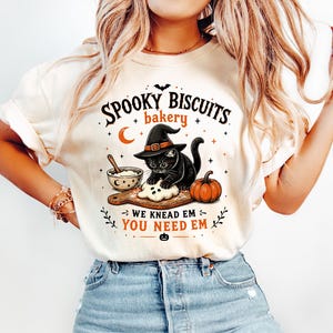 Op de afbeelding: Een cr&egrave;mekleurig t-shirt met een afbeelding van een zwarte kat met een heksenhoed die deeg kneedt op een houten plank naast een kom en een pompoen. De tekst luidt: SPOOKY BISCUITS bakery WE KNEAD EM YOU NEED EM, met kleine sterren, een maan en een vleermuis.