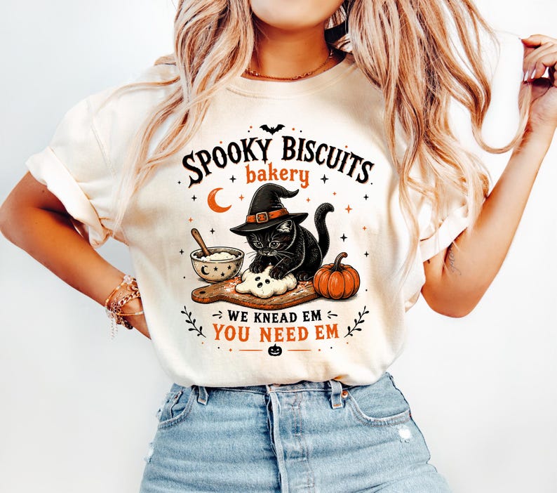 Op de afbeelding: Een cr&egrave;mekleurig t-shirt met een afbeelding van een zwarte kat met een heksenhoed die deeg kneedt op een houten plank naast een kom en een pompoen. De tekst luidt: SPOOKY BISCUITS bakery WE KNEAD EM YOU NEED EM, met kleine sterren, een maan en een vleermuis.