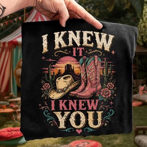 Puede incluir: Una camiseta negra con un estampado gr&aacute;fico de un paisaje des&eacute;rtico con un sombrero de vaquero, botas de vaquero rosas, cactus y rosas. El texto "I KNEW IT" aparece sobre la escena y "I KNEW YOU" est&aacute; impreso debajo en una fuente estilo vintage.