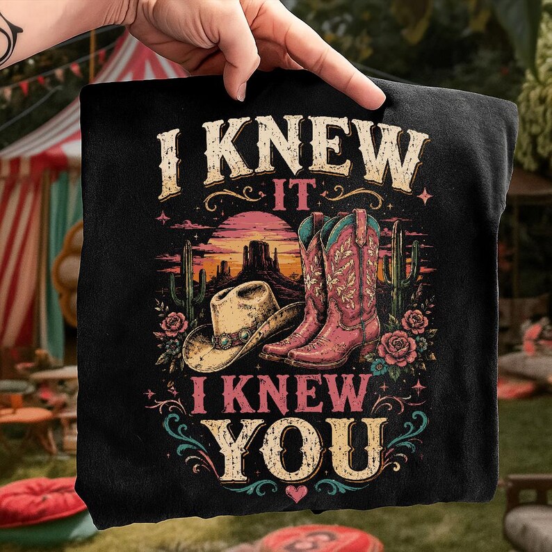 Puede incluir: Una camiseta negra con un estampado gr&aacute;fico de un paisaje des&eacute;rtico con un sombrero de vaquero, botas de vaquero rosas, cactus y rosas. El texto "I KNEW IT" aparece sobre la escena y "I KNEW YOU" est&aacute; impreso debajo en una fuente estilo vintage.