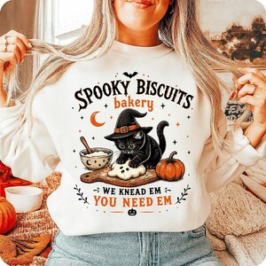 May include: A white crewneck sweatshirt featuring a graphic of a black cat wearing a witch hat, kneading dough on a wooden board. The text reads "SPOOKY BISCUITS bakery" and "WE KNEAD EM YOU NEED EM" with illustrations of a bowl, a pumpkin, and stars.