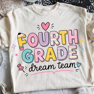 May include: A cream-colored t-shirt featuring a colorful graphic with the text "FOURTH GRADE dream team". The design includes a pencil-shaped letter F, a pink heart, a pushpin, and various stars in shades of pink, yellow, blue, and purple.