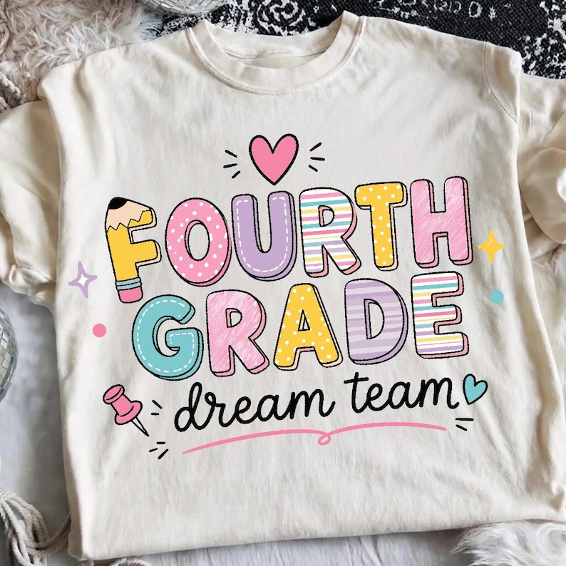 May include: A cream-colored t-shirt featuring a colorful graphic with the text "FOURTH GRADE dream team". The design includes a pencil-shaped letter F, a pink heart, a pushpin, and various stars in shades of pink, yellow, blue, and purple.
