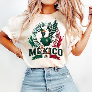 May include: A cream-colored t-shirt featuring a graphic of a white duck wearing a green soccer jersey. The design includes a circular emblem, a soccer ball, and the word MEXICO in bold green letters, accented with red and green brushstroke details.
