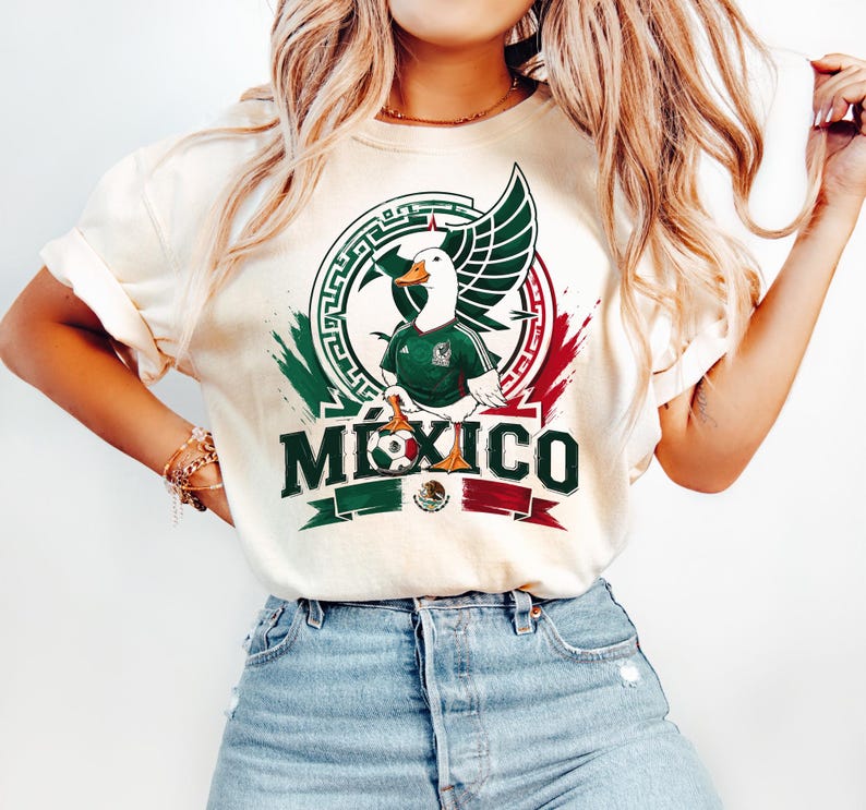 May include: A cream-colored t-shirt featuring a graphic of a white duck wearing a green soccer jersey. The design includes a circular emblem, a soccer ball, and the word MEXICO in bold green letters, accented with red and green brushstroke details.