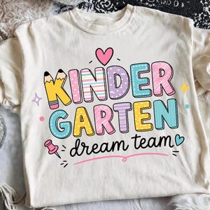 May include: A cream-colored t-shirt featuring the text KINDER GARTEN in large, multi-colored, patterned block letters, with the words dream team in black script below. The design includes a pencil, a pink heart, a pushpin, and small stars.