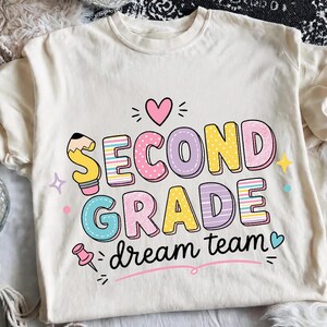 May include: A cream-colored t-shirt featuring the text SECOND GRADE dream team in colorful, patterned bubble letters. The design includes a pencil, a pink heart, a pushpin, and small stars in shades of pink, yellow, blue, and purple.