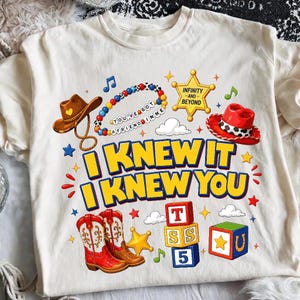May include: A cream-coloured t-shirt featuring a graphic print with a brown cowboy hat, a beaded bracelet with the text YOU'VE GOT A FRIEND IN ME, a yellow sheriff star, a red cowboy hat, and the text I KNEW IT I KNEW YOU above red cowboy boots.