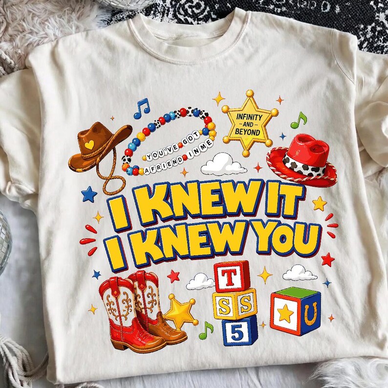 May include: A cream-coloured t-shirt featuring a graphic print with a brown cowboy hat, a beaded bracelet with the text YOU'VE GOT A FRIEND IN ME, a yellow sheriff star, a red cowboy hat, and the text I KNEW IT I KNEW YOU above red cowboy boots.