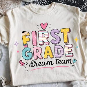May include: A cream-colored t-shirt featuring the text FIRST GRADE in colorful, patterned block letters, with the words dream team written below in black script. The design includes a pencil, a pink heart, a pushpin, and small stars.