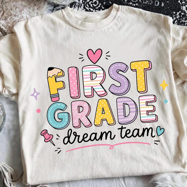 May include: A cream-colored t-shirt featuring the text FIRST GRADE in colorful, patterned block letters, with the words dream team written below in black script. The design includes a pencil, a pink heart, a pushpin, and small stars.