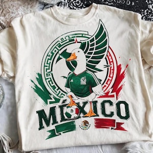 May include: A cream-colored t-shirt featuring a graphic of a white duck wearing a green soccer jersey. The duck stands on a soccer ball with the text M&Eacute;XICO below. The design includes a circular emblem with green and red brushstroke accents.
