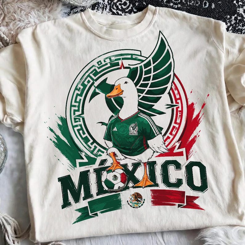 May include: A cream-colored t-shirt featuring a graphic of a white duck wearing a green soccer jersey. The duck stands on a soccer ball with the text M&Eacute;XICO below. The design includes a circular emblem with green and red brushstroke accents.