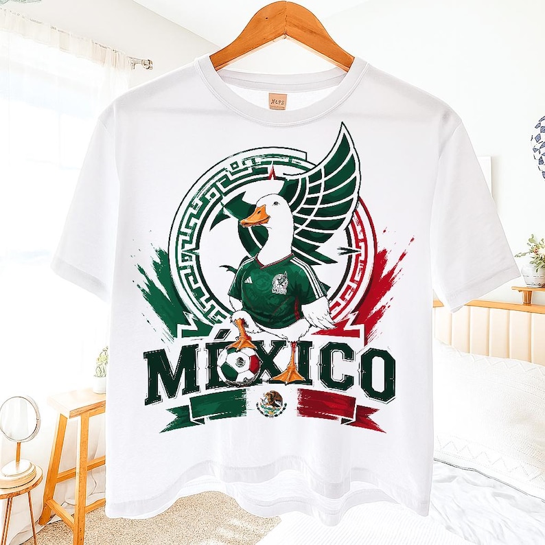 May include: A white short-sleeve t-shirt featuring a graphic of a white duck wearing a green soccer jersey. The duck stands over a soccer ball with the word MEXICO printed in large dark green letters below, accented by red and green brush strokes.