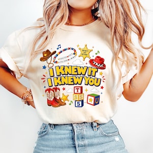 May include: A cream-coloured t-shirt featuring a graphic print with the text "I KNEW IT I KNEW YOU". The design includes a brown cowboy hat, a beaded necklace, a yellow sheriff badge, a red cowboy hat, red boots, and colourful alphabet blocks.