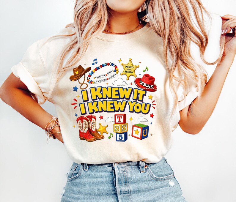 May include: A cream-coloured t-shirt featuring a graphic print with the text "I KNEW IT I KNEW YOU". The design includes a brown cowboy hat, a beaded necklace, a yellow sheriff badge, a red cowboy hat, red boots, and colourful alphabet blocks.