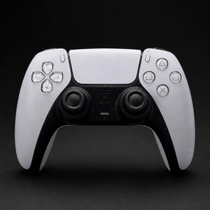 Könnte beinhalten: Ein weiß-schwarzer kabelloser Videospiel-Controller mit einem zentralen Touchpad, zwei schwarzen Analogsticks, einem Steuerkreuz mit vier transparenten Tasten sowie vier Aktionstasten auf der rechten Seite mit Dreieck-, Kreis-, Quadrat- und Kreuzsymbolen.