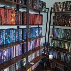 May include: A room with bookshelves packed with books. The books display a range of colours and patterns on their spines. The shelves are made of dark wood and supported by black metal frames, creating a visually appealing display.