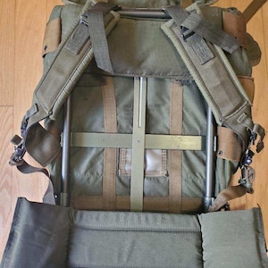 May include: An olive green external frame backpack featuring a metal frame structure, adjustable padded shoulder straps, and a wide padded waist belt. The pack includes brown webbing straps, a top flap, and a clear plastic identification window.