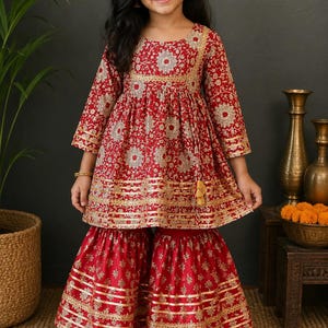 May include: A red and gold embroidered dress and matching pants set for a young girl. The outfit features a floral pattern and gold trim. The dress has long sleeves and a flared skirt, while the pants are wide-legged. The girl is wearing gold shoes.