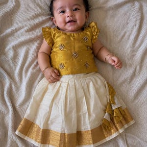 May include: A two-piece outfit consisting of a mustard yellow sleeveless top with ruffled shoulders and floral bead embellishments, paired with an off-white pleated skirt featuring a wide gold metallic border and a vertical ruffled side accent.