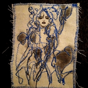 May include: A rectangular fabric patch with a beige base featuring a dark brown ink illustration of a figure with long hair. The design is outlined with vibrant blue zigzag embroidery stitching along the edges and throughout the central image.