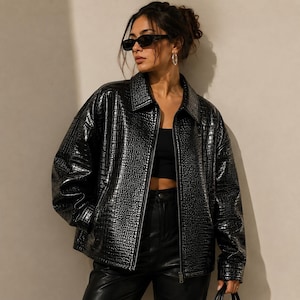 Könnte beinhalten: Eine schwarze, übergroße Jacke mit Krokodilhautstruktur. Die Jacke hat einen Kragen, einen Reißverschluss und lange Ärmel. Das Model trägt eine schwarze Sonnenbrille und Creolen. Die Jacke wird mit einer schwarzen Hose kombiniert.