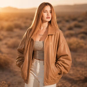 Könnte beinhalten: Eine beige Bomberjacke aus Kunstleder mit Kragen, langen Ärmeln und einem durchgehenden Frontreißverschluss. Die Jacke wird über einem grauen trägerlosen Oberteil und einer weißen Hose getragen. Die Jacke hat eine glatte Oberfläche und gerippte Bündchen.