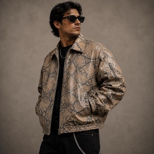 May include: A tan and brown snakeskin-patterned jacket with a zipper closure and a small pocket on the left sleeve. The jacket has a collared neckline and is worn with black pants and sunglasses.