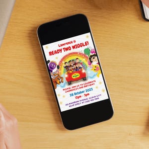 May include: A smartphone displaying a digital birthday invitation for Lawrence. The design features The Wiggles characters in a red car under a rainbow, with text reading Lawrence is READY TWO WIGGLE, 28 October 2023, 12pm-3pm, and address details.