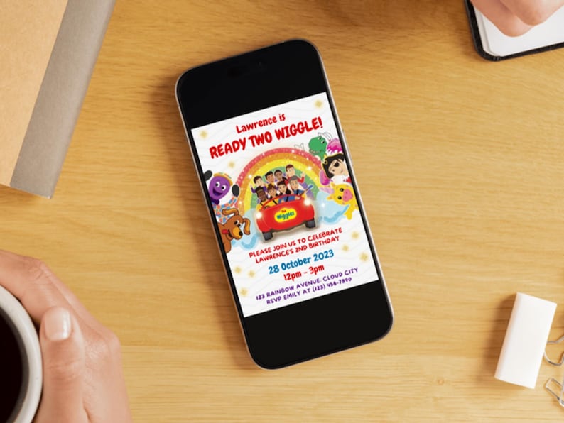 May include: A smartphone displaying a digital birthday invitation for Lawrence. The design features The Wiggles characters in a red car under a rainbow, with text reading Lawrence is READY TWO WIGGLE, 28 October 2023, 12pm-3pm, and address details.