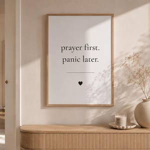 Könnte beinhalten: Gerahmter Druck mit dem Text "prayer first. panic later." in minimalistischem Stil. Unter dem Text befindet sich eine horizontale Linie und ein schwarzes Herz. Der Druck befindet sich in einem hellen Holzrahmen und wird an einer hellen Wand angezeigt.