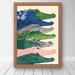 May include: A framed art print featuring a vertical stack of seven alligator heads in various colours and patterns, including solid green, dark blue, spotted, and pink, all rendered with detailed line work against a light cream background.