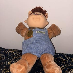 Puede incluir: Muñeca de gato de peluche con cara de color canela claro, pelaje marrón y peto vaquero azul. La muñeca tiene pelo castaño y un collar marrón con una medalla dorada. El peto tiene un pequeño parche.