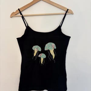 May include: Black camisole top with thin straps, featuring three jellyfish appliqués. The jellyfish have light blue floral patterned caps and yellow tendrils. The top is displayed on a wooden hanger, ready to wear.