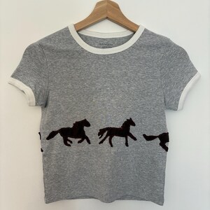 May include: A grey t-shirt with white trim around the neckline and sleeves. The shirt has a design of dark brown horse silhouettes across the front. The t-shirt is hanging on a wooden hanger.