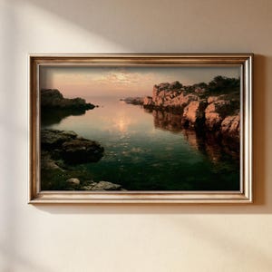 May include: A rectangular landscape painting in a metallic gold frame depicting a calm coastal inlet at sunset. The scene features dark rocky cliffs on both sides of the water, with the soft orange and pink light of the setting sun reflecting on the surface.