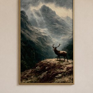 Könnte beinhalten: Ein gerahmter Kunstdruck, der einen majestätischen Hirsch auf einem felsigen, grasbewachsenen Hügel zeigt. Der Hintergrund zeigt eine dramatische, stimmungsvolle Berglandschaft mit tiefen Schatten, nebligen Tälern und durch dunkle Wolken brechenden Lichtstrahlen.