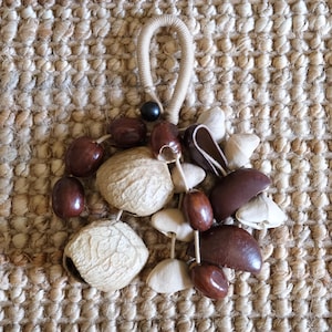 May include: A handcrafted, natural-toned ornament featuring a variety of seeds and nuts, including brown, beige, and white elements. The ornament is suspended by a woven cord and a black bead. The background is a textured, woven surface.