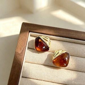 May include: A pair of stud earrings featuring a smooth, rounded triangular amber-colored resin stone set against a polished gold-tone metal accent with three distinct diagonal ridges on the upper corner of each piece.