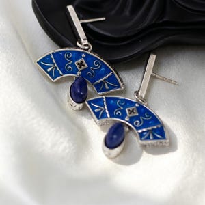 Puede incluir: Un par de pendientes de botón plateados con un diseño en forma de abanico, esmalte azul vibrante, filigranas plateadas y un recorte central en forma de rombo. Cada pendiente está decorado con una piedra azul lisa de forma ovalada.