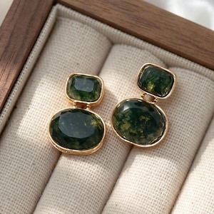 May include: A pair of gold-toned earrings featuring two faceted dark green moss agate stones each. The top stone is rectangular with rounded corners, while the bottom stone is larger and oval-shaped. Both stones are set in gold metal frames.