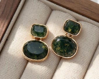 May include: A pair of gold-toned earrings featuring two faceted dark green moss agate stones each. The top stone is rectangular with rounded corners, while the bottom stone is larger and oval-shaped. Both stones are set in gold metal frames.