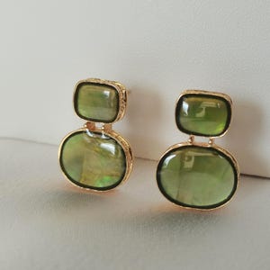 May include: A pair of gold-toned earrings featuring two stacked, smooth, olive-green stones. The top stone is rectangular with rounded corners, while the bottom stone is oval. Each stone is encased in a textured gold-colored metal frame.