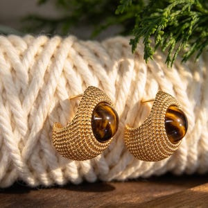 May include: A pair of gold-toned hoop earrings featuring a textured, beaded surface and a central, smooth, round tiger's eye gemstone. The earrings have a wide, semi-circular shape with a polished finish and a post-back closure mechanism.