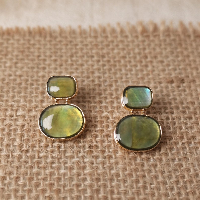 May include: A pair of gold-toned earrings featuring two stacked, smooth, olive-green oval stones. The smaller stone sits above the larger, rounded stone, both encased in a thin gold metal frame with a polished, reflective surface finish.