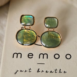 May include: A pair of gold-toned earrings featuring two stacked, smooth, iridescent green stones in rectangular and oval shapes. The earrings are displayed on a cream-colored card with the black text memoo just breathe.