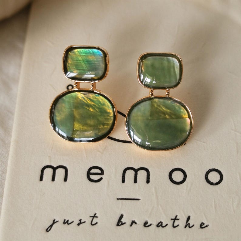 May include: A pair of gold-toned earrings featuring two stacked, smooth, iridescent green stones in rectangular and oval shapes. The earrings are displayed on a cream-colored card with the black text memoo just breathe.